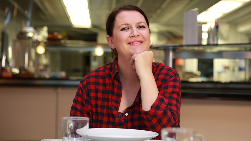 Titelbild des Podcasts "Das geht auf mich" mit Maja Herzbach, die auf diesem Foto vor einem leeren Teller in der Kantine des NDR sitzt und in die Kamera lächelt. 